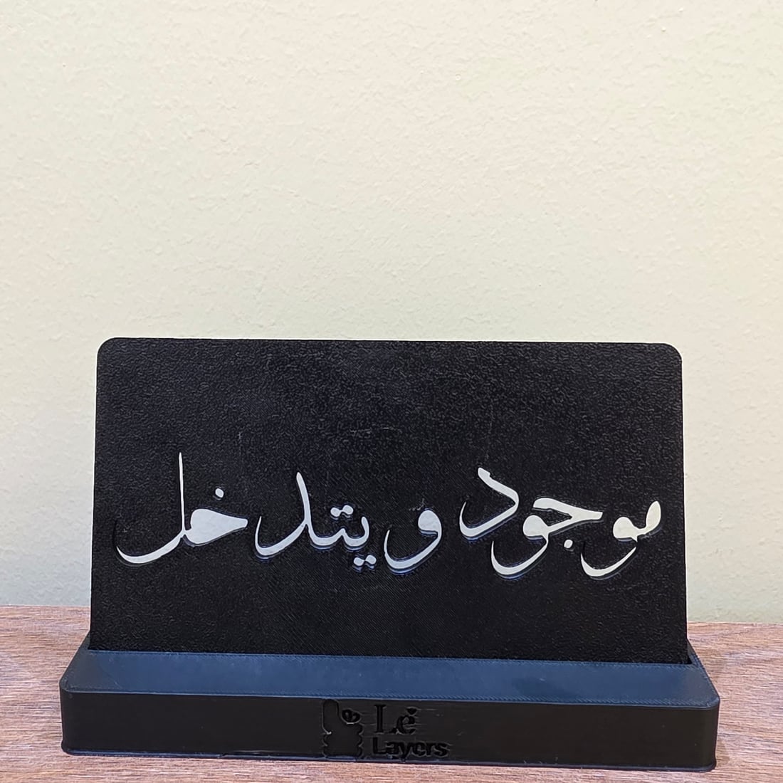 A matte black 3D-printed standing plaque with the Arabic phrase موجود و يتدخل cut through it and backed in gold, on a Le Layers branded base.